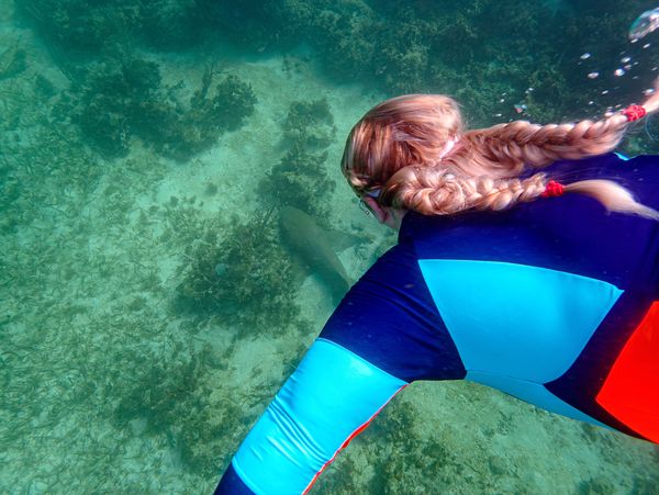 A snorkeler with braided hair swims near a shark in clear water.
