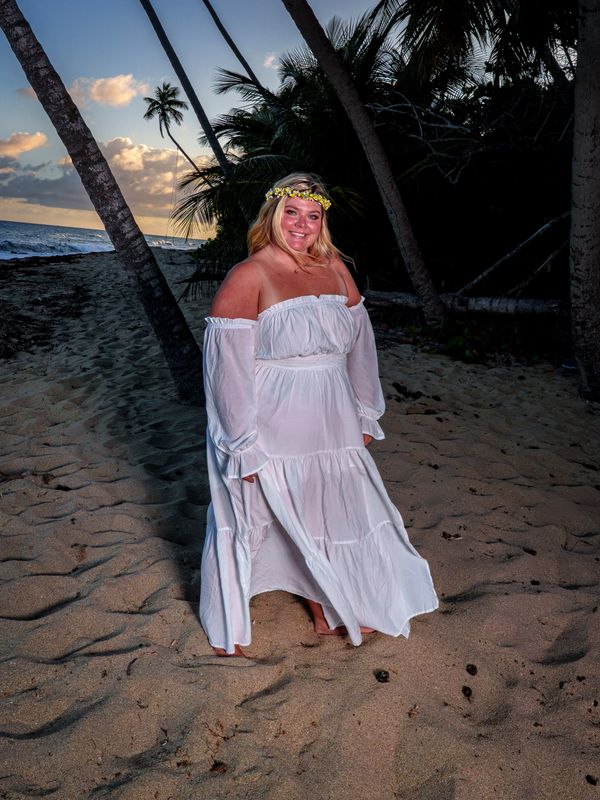 Woman in a flowing white dress and flower crown on a sandy beach at sunset.