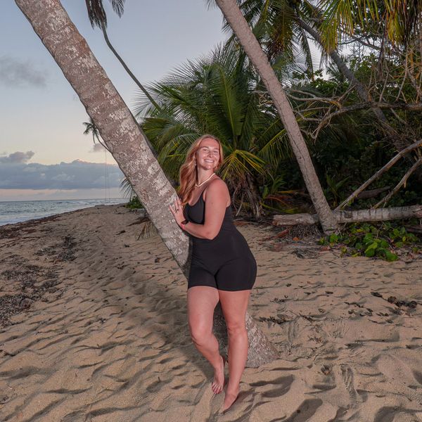Woman in black outfit leaning on a palm tree at the beach during sunset.