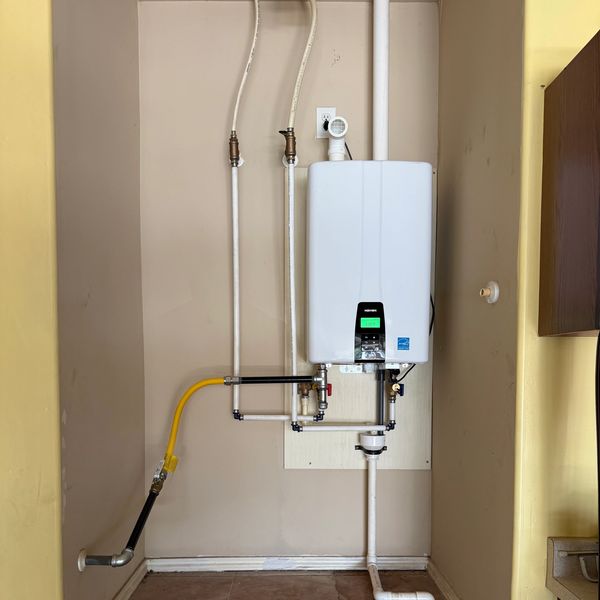 A modern tankless water heater installed on a beige wall with connected pipes.