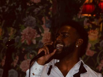 A man passionately singing into a microphone with floral wallpaper in the background.
