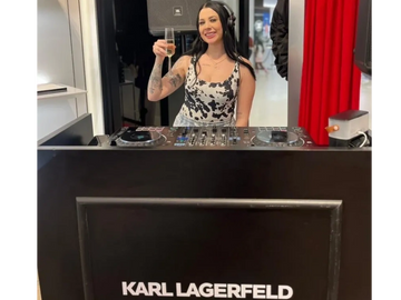 DJ woman raising a glass behind a Karl Lagerfeld-branded DJ booth.