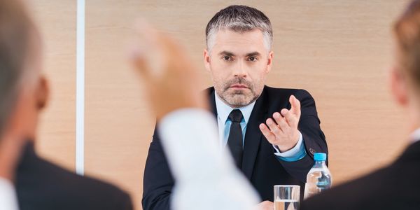 A man in a suit gestures while talking during a meeting.