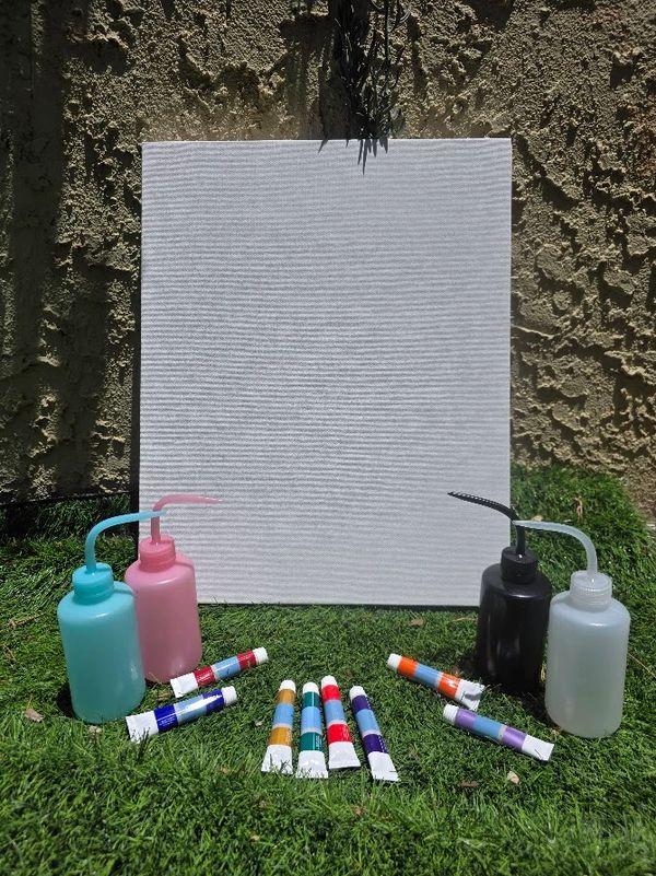 Blank canvas with paint bottles and colorful paint tubes on grass outdoors.