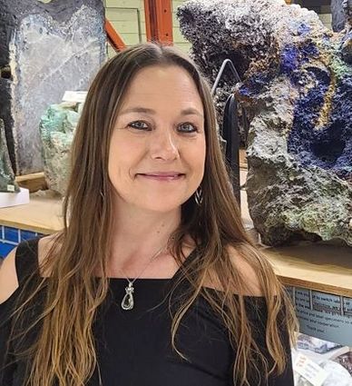 Smiling woman with long hair in a black top, standing indoors near colorful mineral or rock displays.