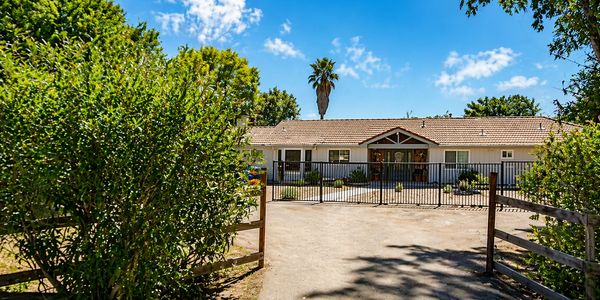 Single-story house with gated entrance surrounded by greenery under a blue sky.