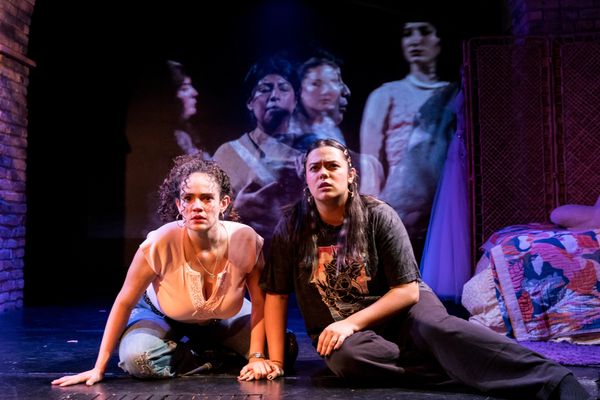 Two women on stage with intense expressions, projected images of faces behind.