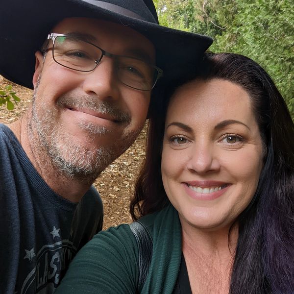 Smiling couple outdoors, close-up selfie with greenery in the background.