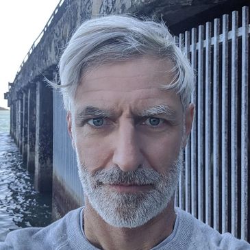A mature man with grey hair and beard standing near a waterfront structure.