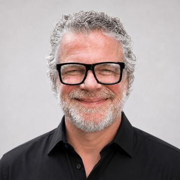 Smiling middle-aged man with curly gray hair and beard wearing black glasses and shirt.