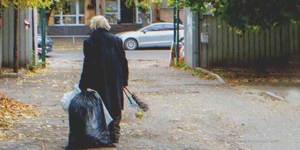 An elderly person walking down a street carrying a large bag and broom.