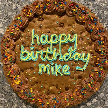 A large chocolate chip cookie cake with chocolate frosting and colorful sprinkles saying 'happy birthday mike'.