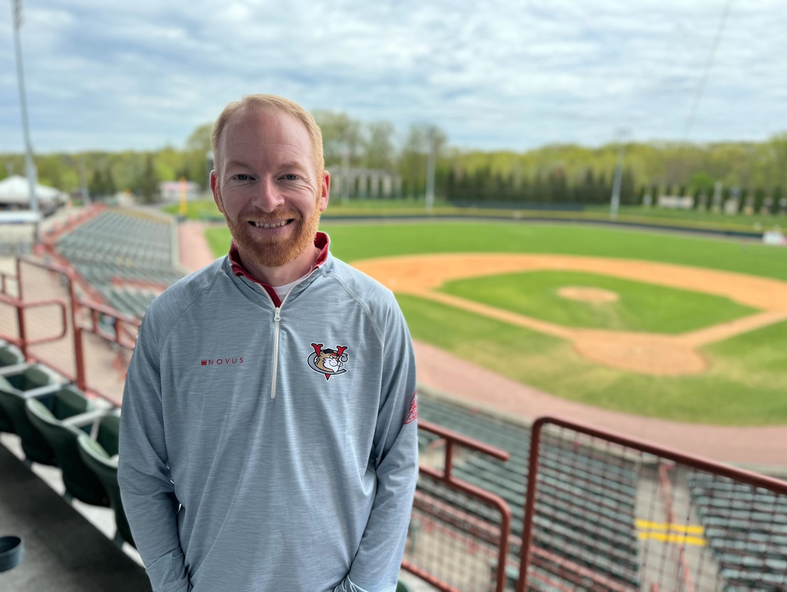 White male wearing a grey pullover sweatshirt poses for a photo in front of empty baseball field. 