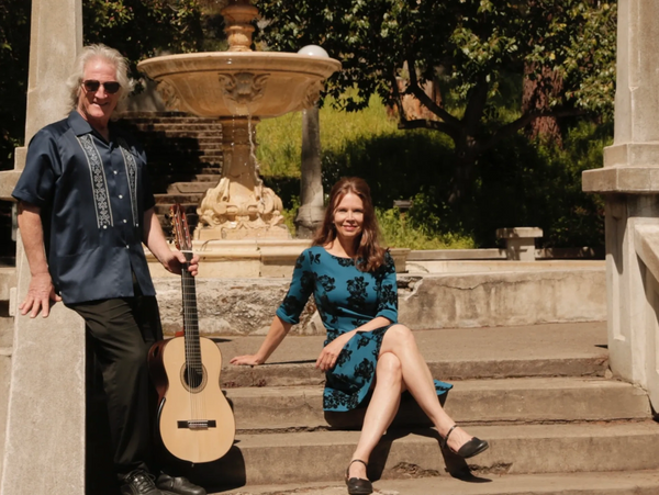 Man with guitar and woman in blue dress posing on stone steps near fountain.
