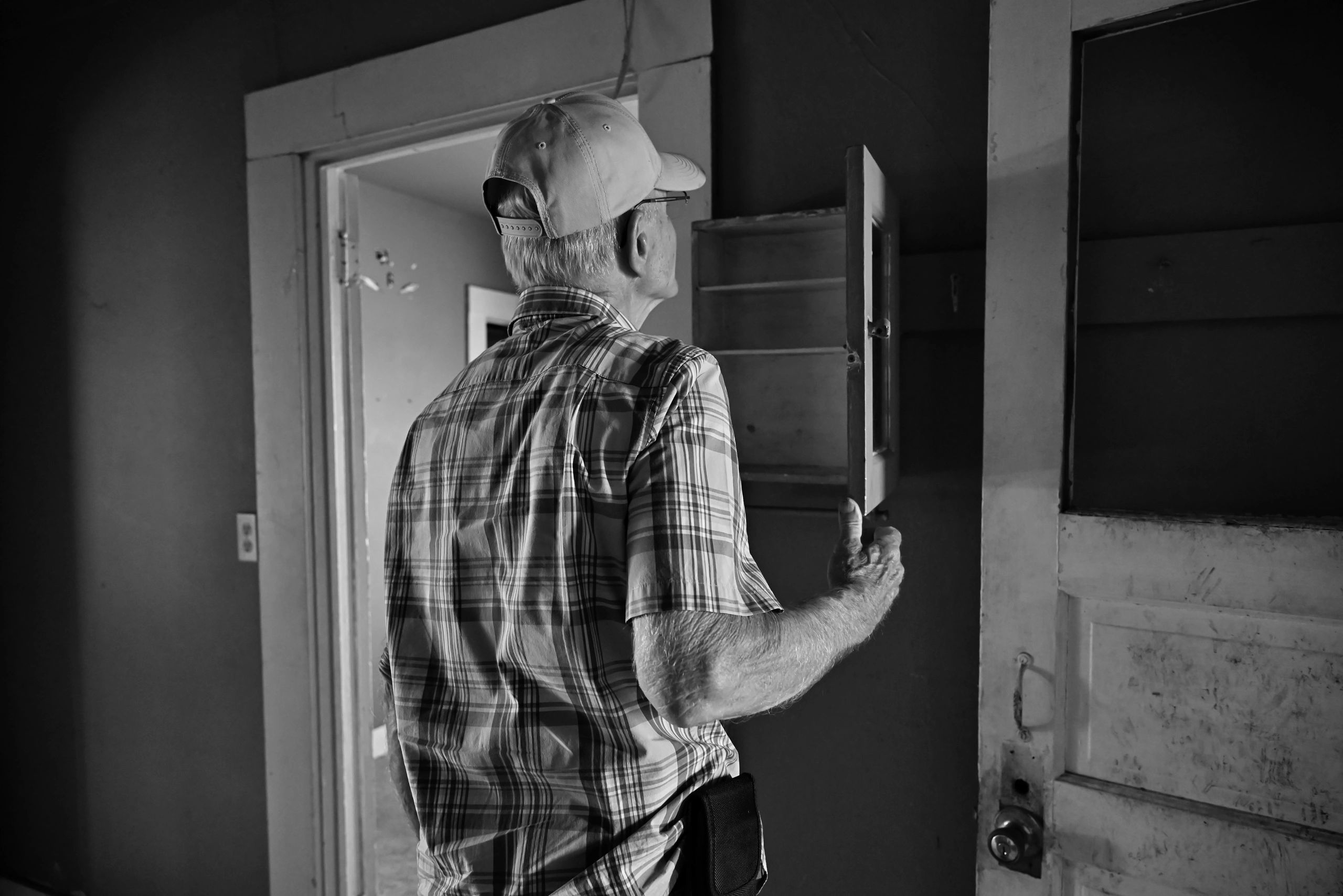 Elderly man in cap opening a small cabinet in an old room.
