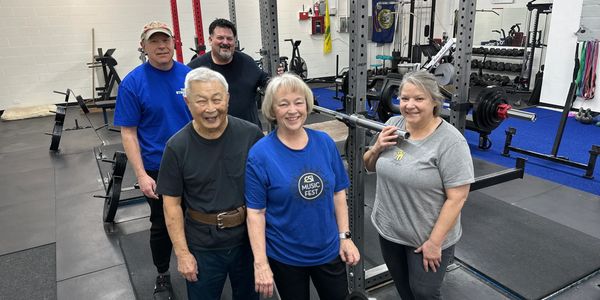 A group of five smiling adults posing in a gym with weightlifting equipment.