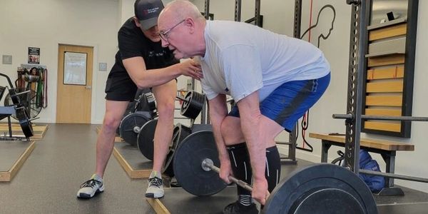 Older man deadlifting with trainer's guidance in a gym.