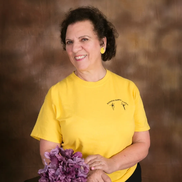 Smiling woman in yellow shirt holding purple flowers against a brown backdrop.