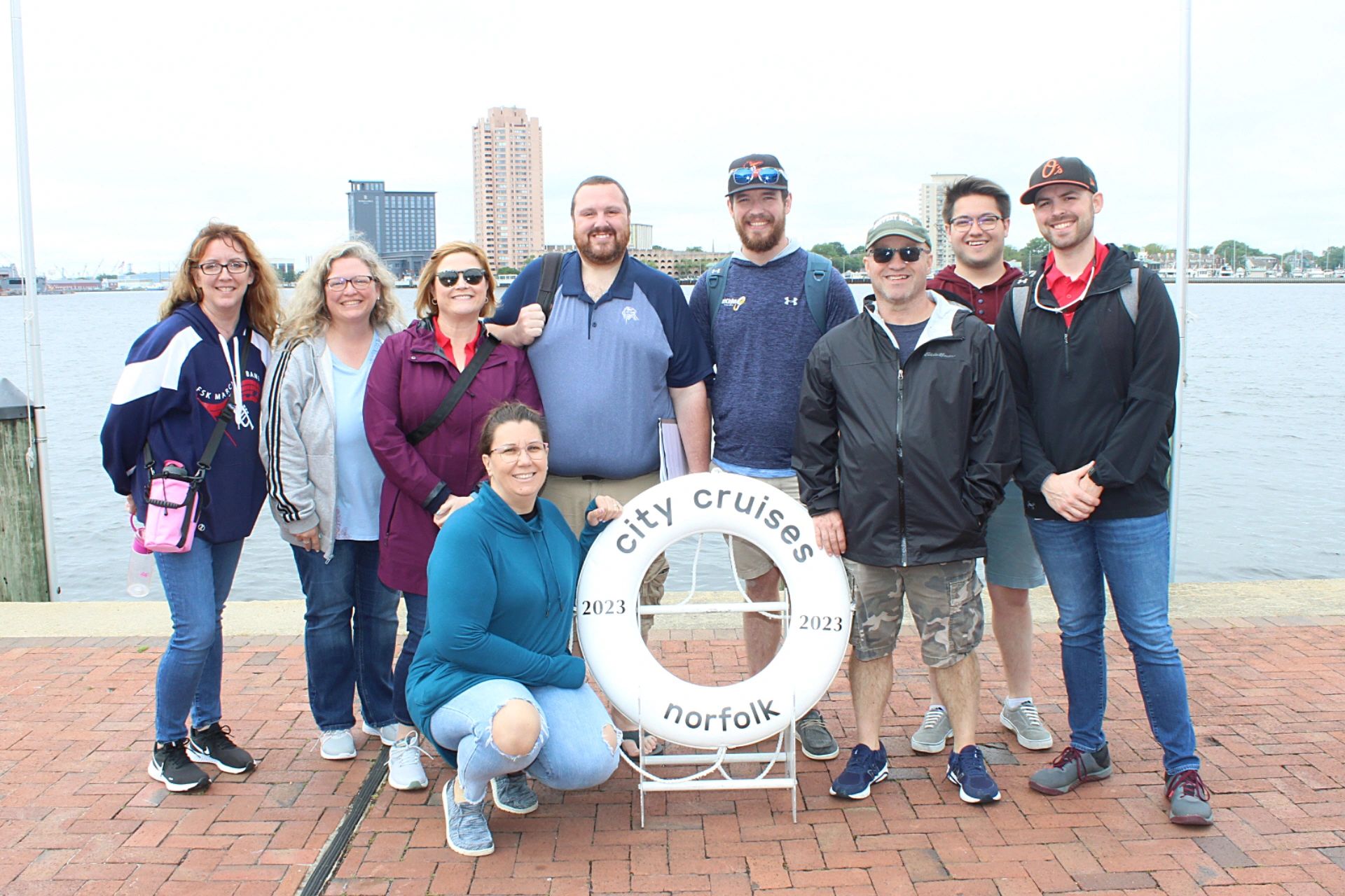 Group of nine people posing by the waterfront with a 'city cruises norfolk' life ring.