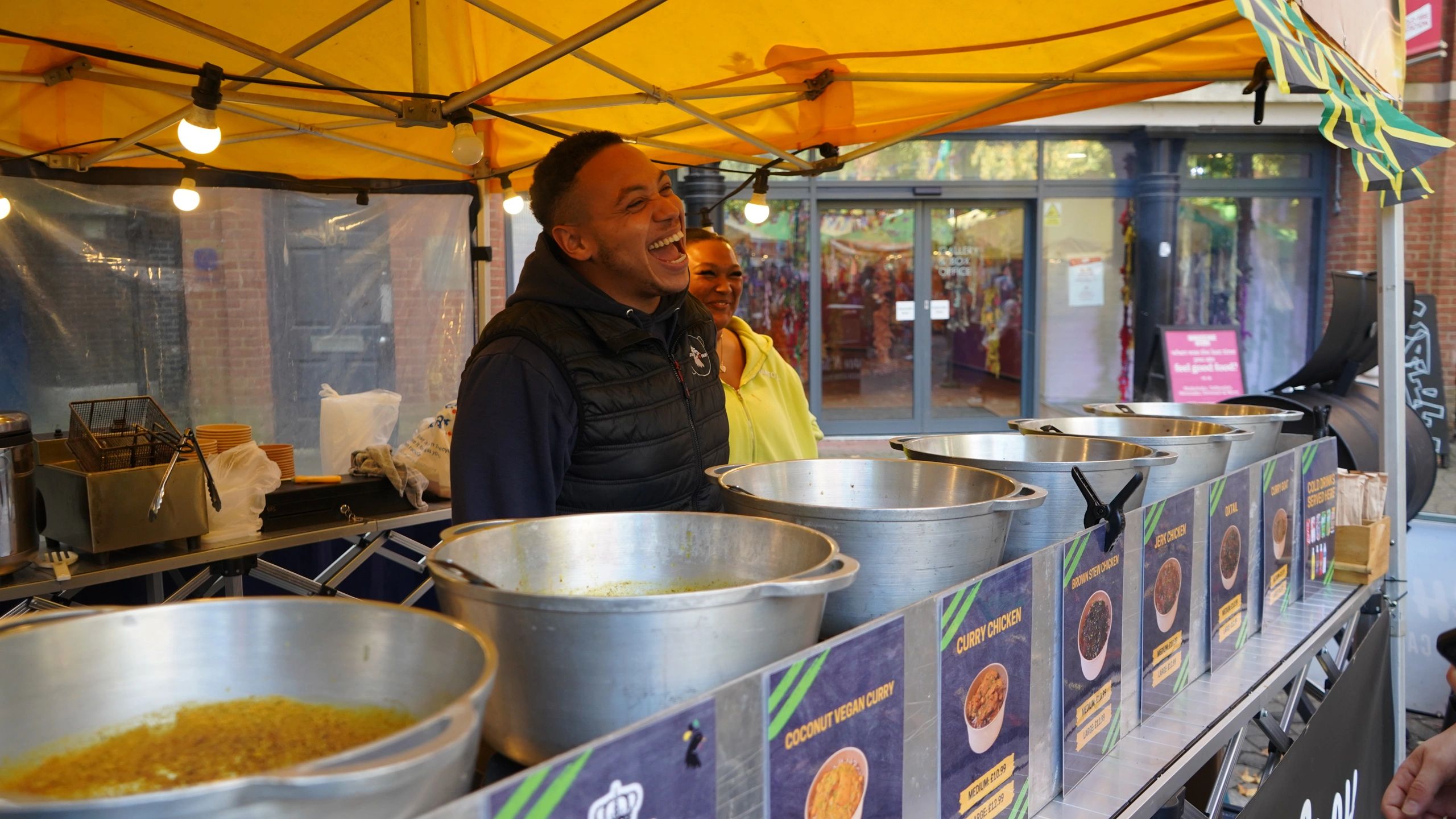 Two people laughing behind a food stall with large pots of curry dishes.