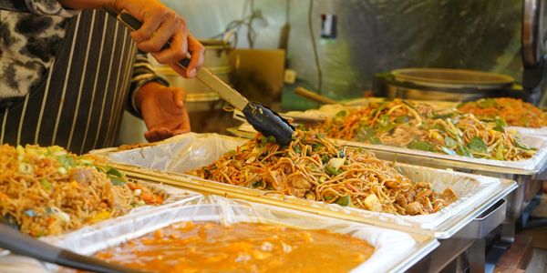 Person serving stir-fried noodles from a buffet tray.