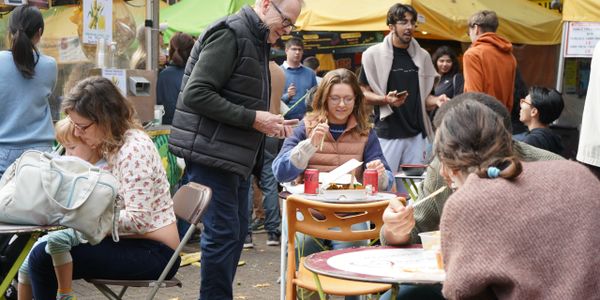 People enjoying food and conversation at an outdoor market with yellow tents.