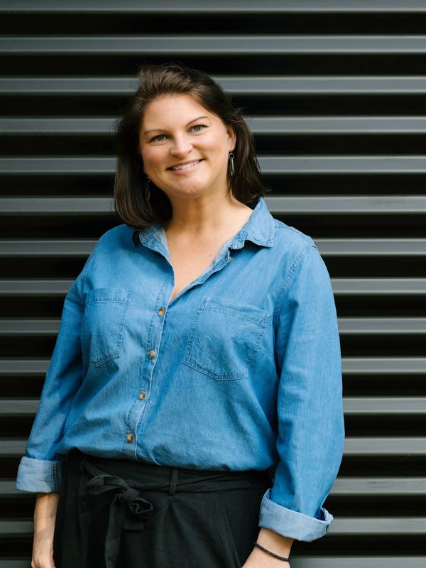 Smiling woman in a denim shirt stands against a striped black background.
