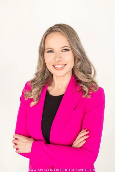 Confident woman in a bright pink blazer smiles with arms crossed against a white background.