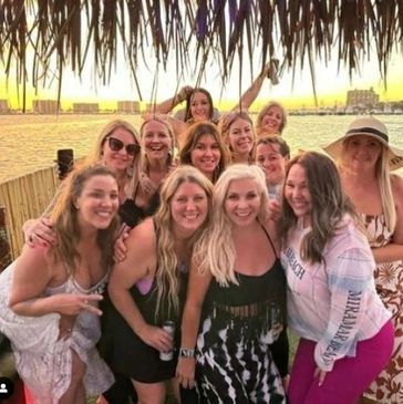 Group of women smiling and posing under a thatched roof by the water at sunset.
