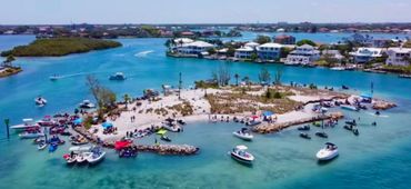 Boats and jet skis gather around a small sandy island near waterfront homes.