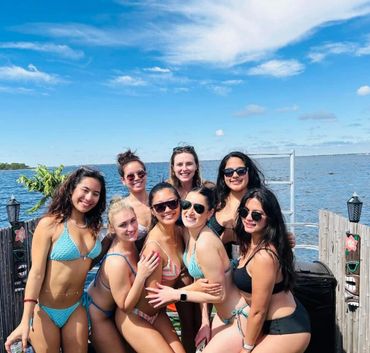 Group of eight women in bikinis posing happily by the water under a bright blue sky.