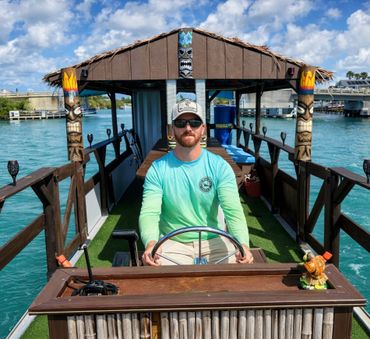 Man steering a tiki-themed boat on clear blue water under a partly cloudy sky.