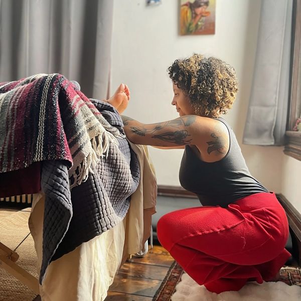 Person with tattoos stretching arms on a bed, wearing red pants and a black top.