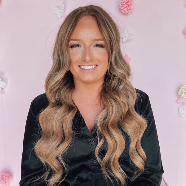 Smiling woman with long wavy blonde hair and black shirt against pink floral background.