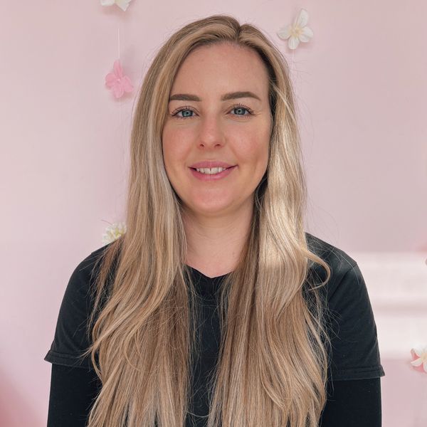 Smiling woman with long blonde hair against a soft pink background with flowers.