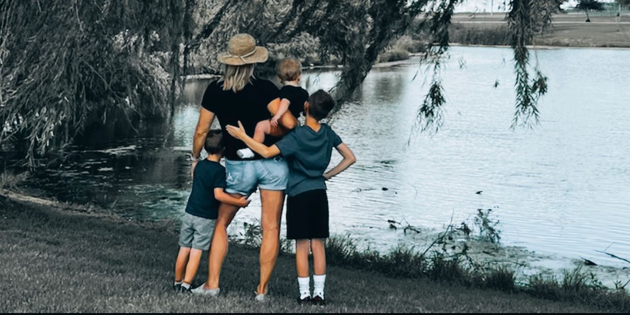 A woman with three children by a serene lake under hanging tree branches.