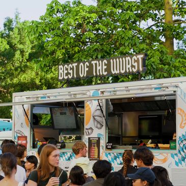 A crowd waiting at a food truck named 'Best of the Wurst' on a sunny day.