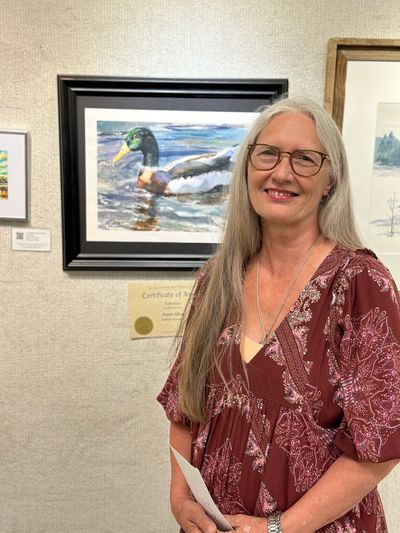 Smiling woman with long gray hair stands beside a framed duck painting in an art gallery.