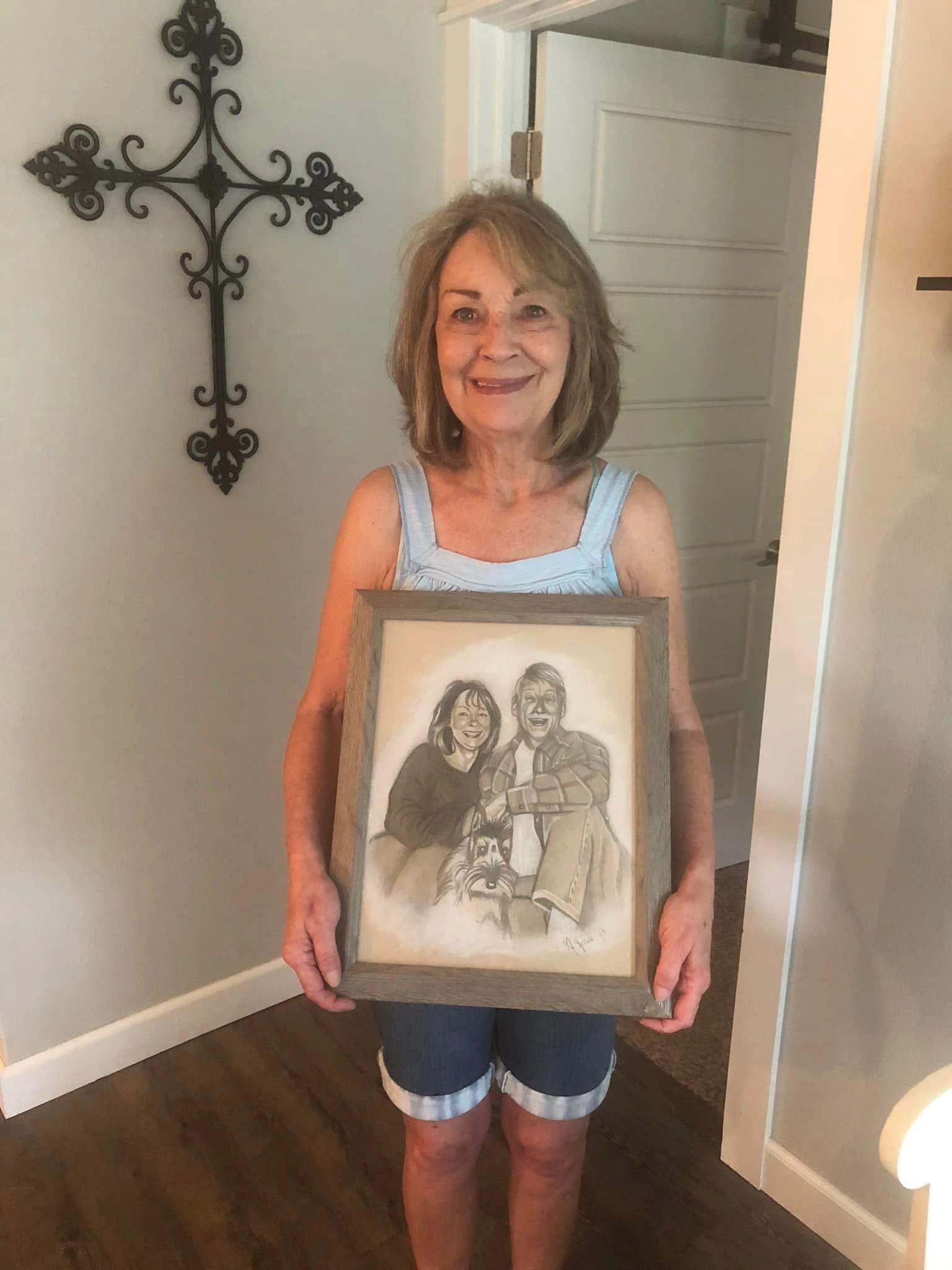 Smiling woman holding a framed drawing of a couple with a dog.
