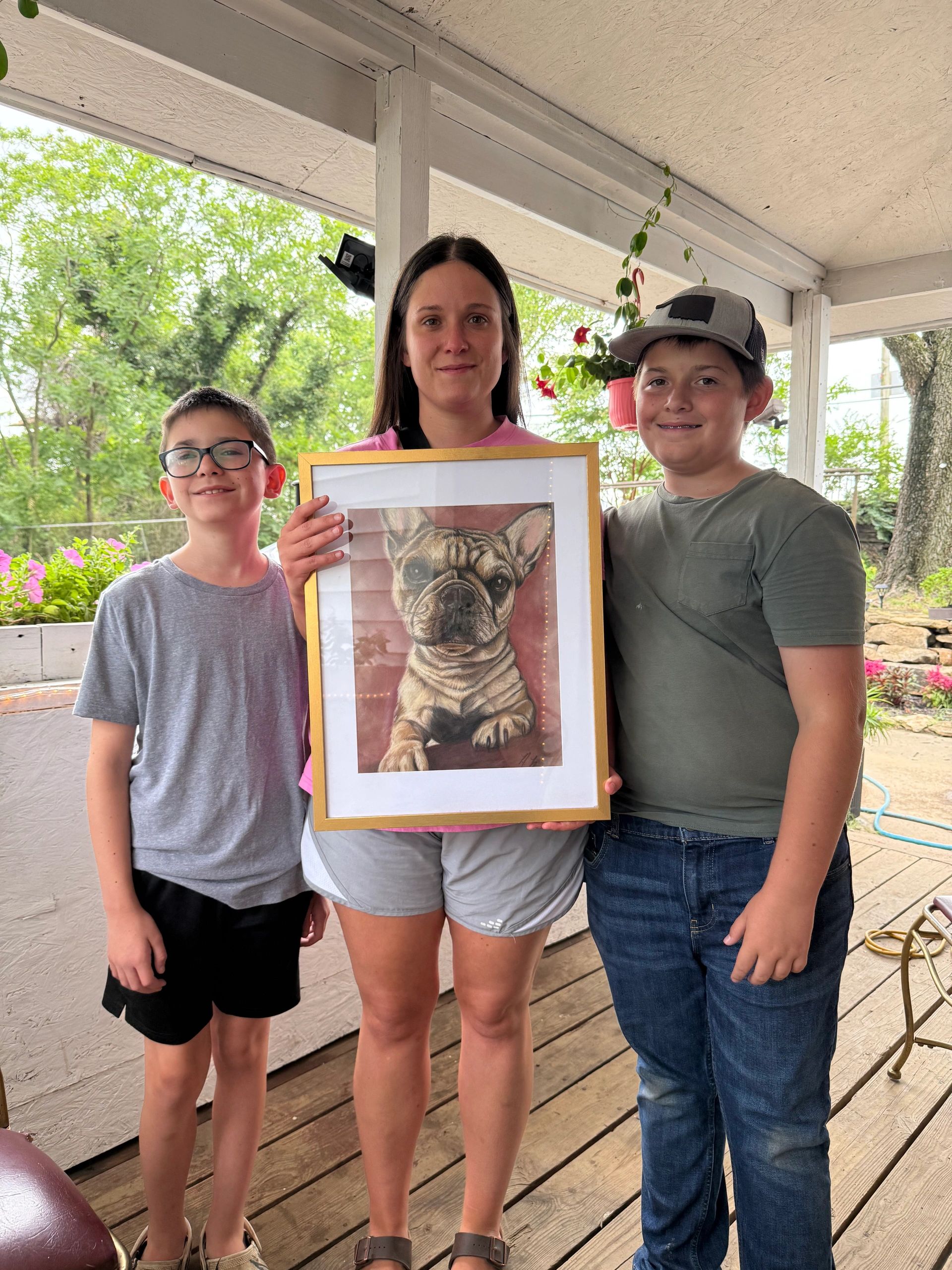 Three people holding a framed drawing of a French Bulldog, smiling outdoors on a porch.