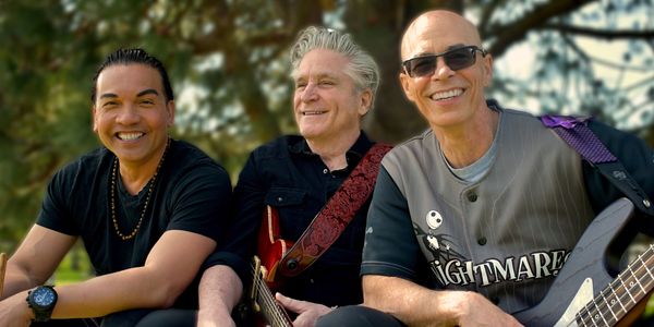 Three smiling musicians outdoors, holding guitars and drumsticks.