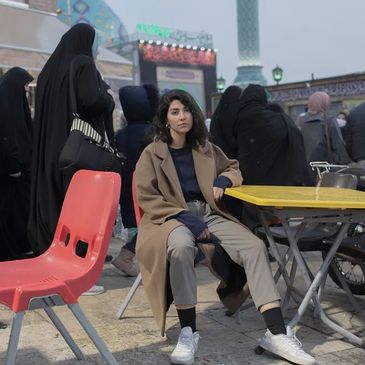A woman in casual attire sits at a yellow table amid people in traditional black clothing.