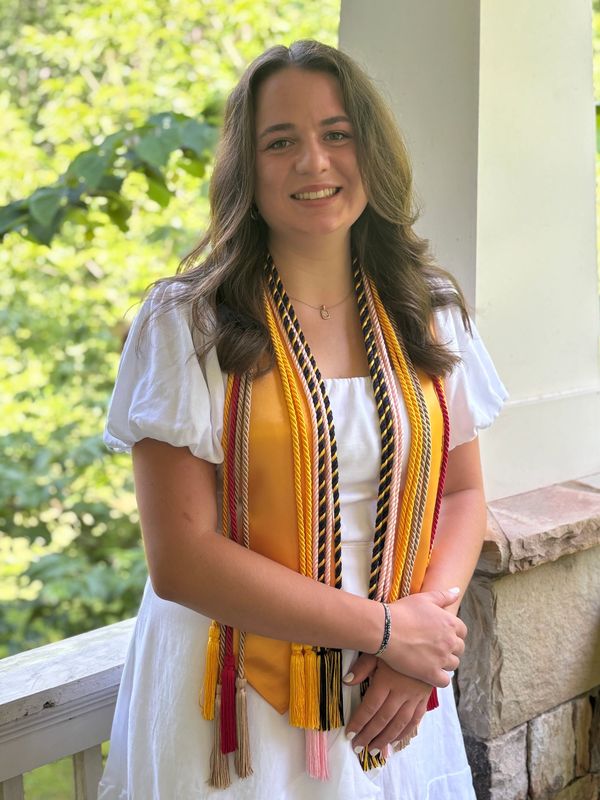 Young woman in a white dress with honor cords, smiling on a porch.