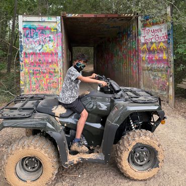 A boy wearing a mask sitting on a muddy ATV near a graffiti-covered metal structure.