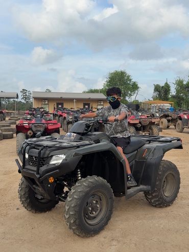 A boy rides a black ATV on a dirt lot with other ATVs in the background.