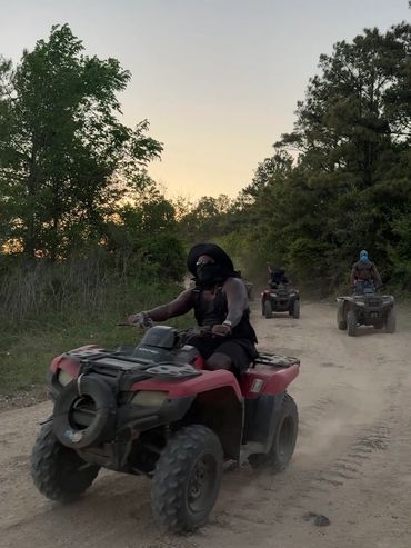 Three people riding ATVs on a dusty trail surrounded by trees at dusk.