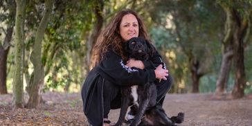 Woman hugging her black dog while crouching on a tree-lined path.