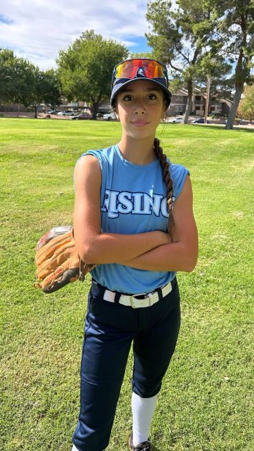 Young female softball player in uniform standing on the field with arms crossed.