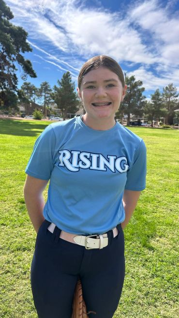 A smiling young athlete in a blue "RISING" shirt stands on a sunny grassy field.
