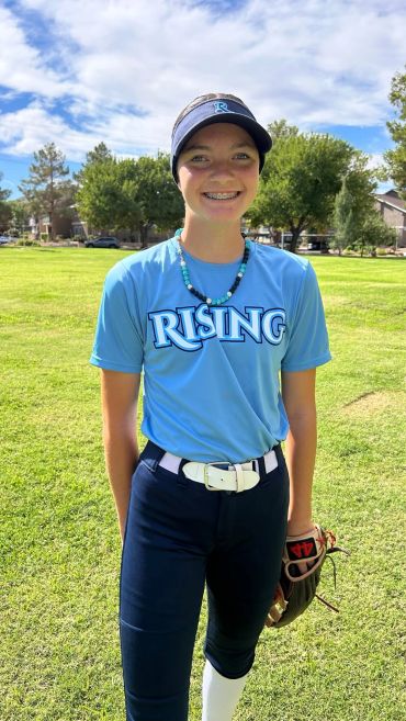 A smiling young baseball player in uniform stands on a sunny field.
